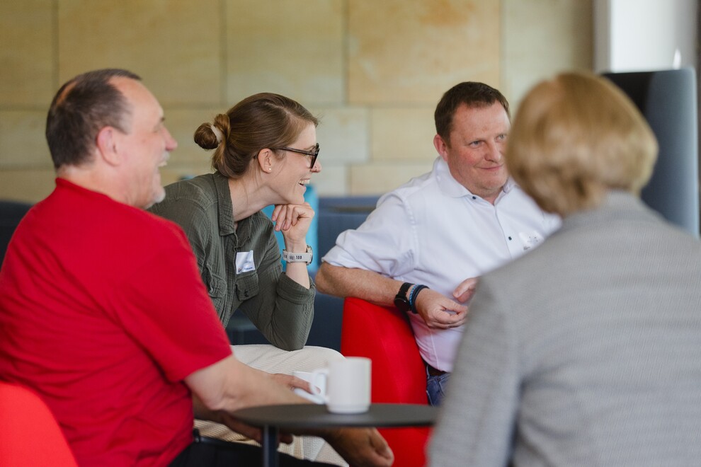 Vier Erwachsene sitzen in gemütlicher Runde um einen kleinen Tisch und unterhalten sich lachend. Zwei Männer und eine Frau sind frontal sichtbar, eine weitere Person ist von hinten zu sehen.