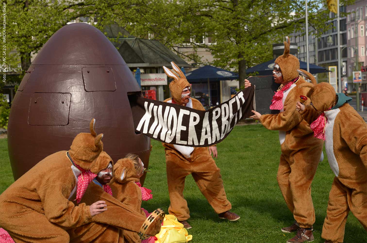 Eine Gruppe von Menschen, verkleidet als Osterhasen, vor einem riesigen schokoladenbraunen  Ei in Form einer Riesen-Handgranate. Einer der Verkleideten zieht aus dem Ei ein Transparent auf dem steht: KINDERARBEIT