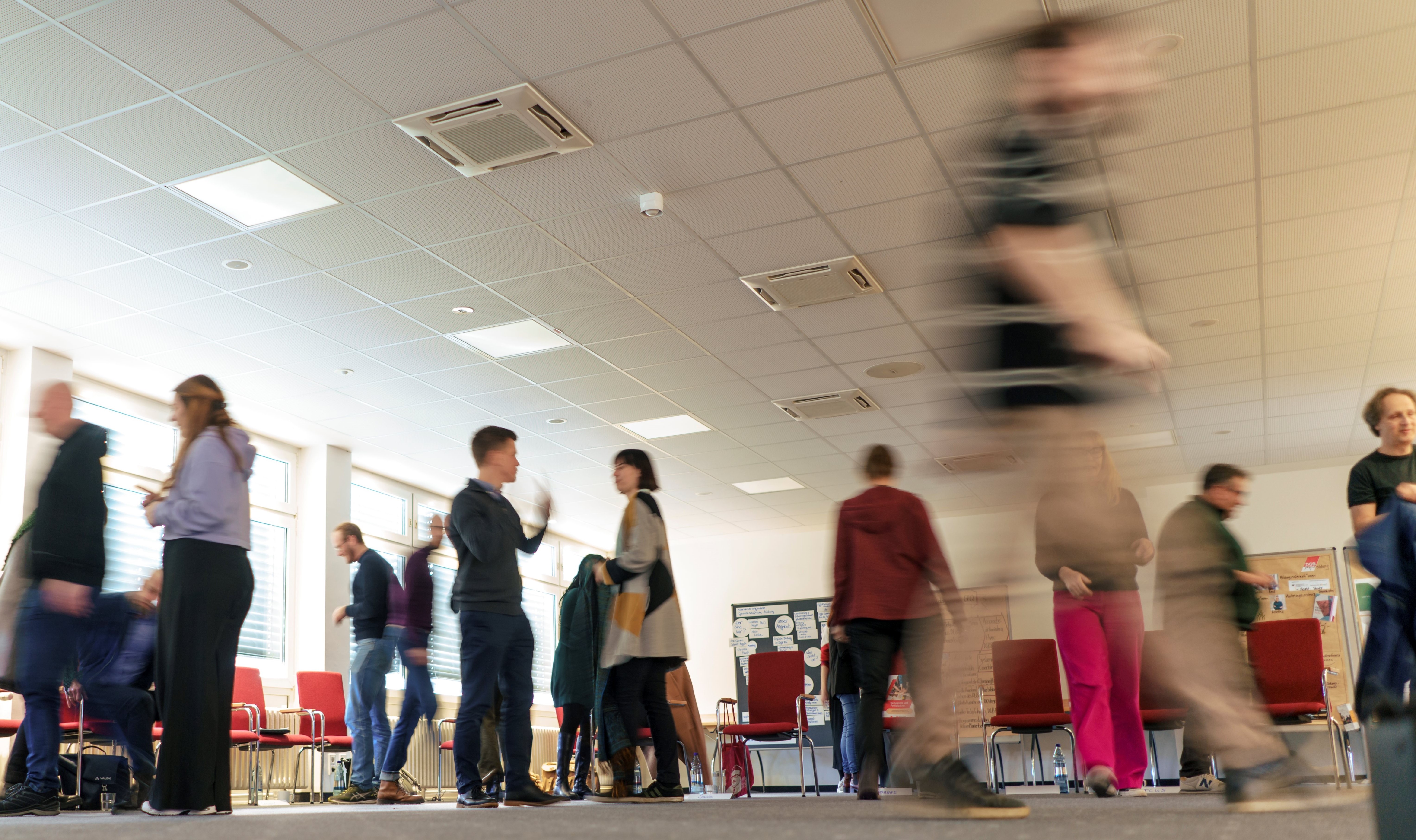 Menschen stehen in einem großen Raum mit roten Stühlen im Kreis, im Hintergrund sind Karten an Wänden für eine Schulung.  