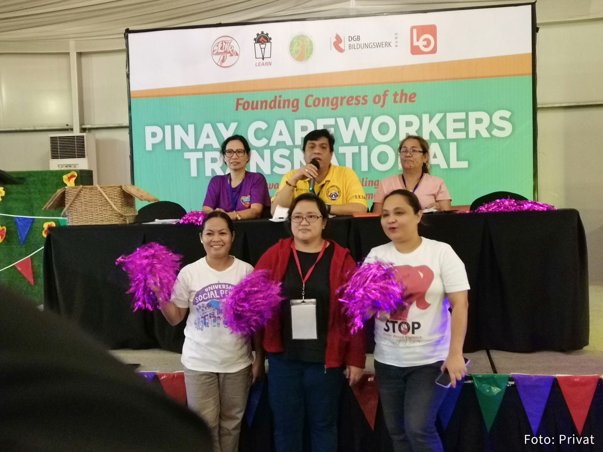 Gewerkschaften sitzen am Podium und stehen davor. Im Hintergrund ist eine große Leinwand auf der steht: Founding Congress of Pin@y Transnational Care Workers