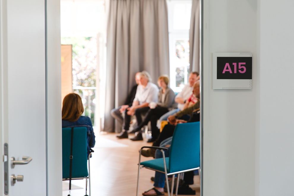 Eine Gruppe von Personen sitzt in einem Raum mit großen Fenstern und grauen Vorhängen. An der Wand hängt ein Schild mit der Aufschrift „A15“ in pinker Schrift.