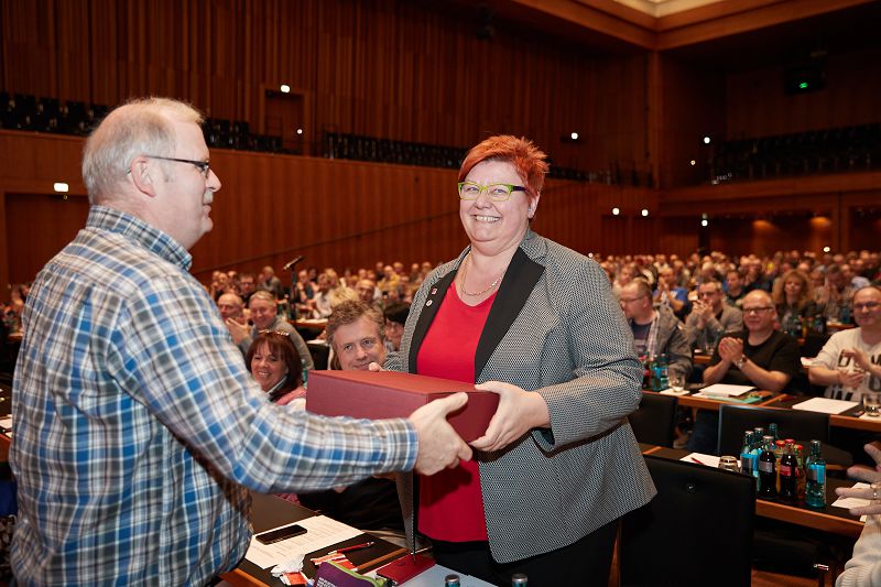 Eine Frau mit rotem Haar und Brille nimmt auf einer Bühne in einem großen Konferenzsaal lächelnd ein Geschenk von einem Mann entgegen. Im Hintergrund applaudiert das Publikum, das an Tischen sitzt.