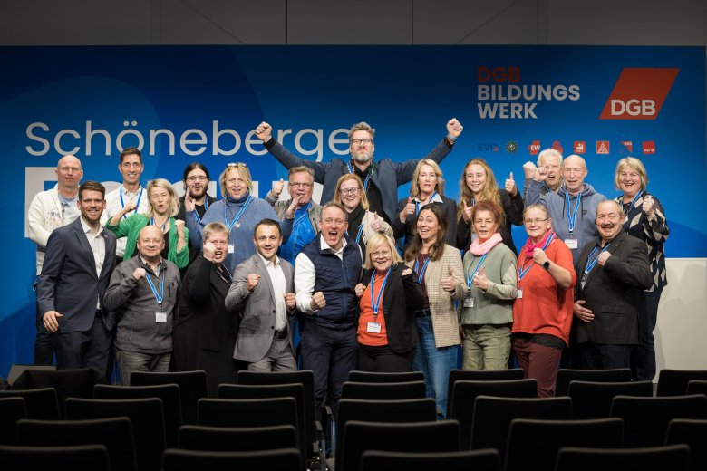 Gruppe reckt die Arme und steht vor einem blauen Rückwand mit dem Schriftzug Schöneberger Forum