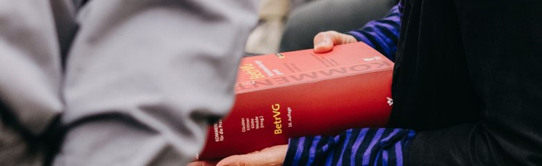Hände halten ein BetrVG-Kommentarbuch, im Vordergrund eine Person mit grauem Hemd.