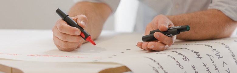 Eine Person schreibt mit einem roten und einem schwarzen Marker auf ein großes Blatt Papier, das auf einem Tisch liegt. Der Text auf dem Papier ist teilweise sichtbar und enthält handgeschriebene Notizen.