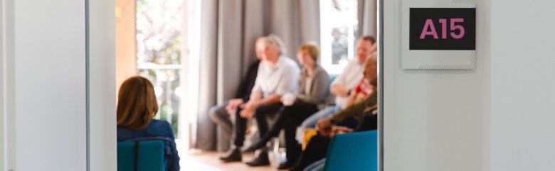 Eine Gruppe von Personen sitzt in einem Raum mit großen Fenstern und grauen Vorhängen. An der Wand hängt ein Schild mit der Aufschrift „A15“ in pinker Schrift.