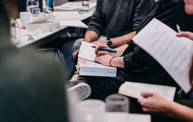 Hände halten ein Arbeitsgesetzbuch während eines Seminars, auf dem Tisch sind Notizen sichtbar.