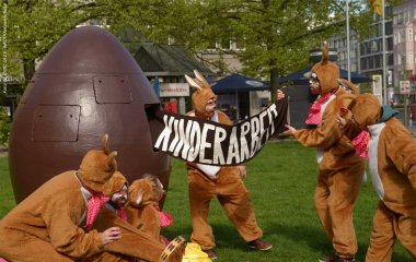 Als Osterhasen verkleidete Personen ziehen einen Banner mit der Aufschrift "Kinderarbeit" aus einem menschengroßen Schokoei.